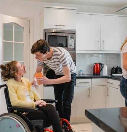 Teenage boy is assisting his disabled sister with having a drink in the kitchen of their home while their parents make lunch.
